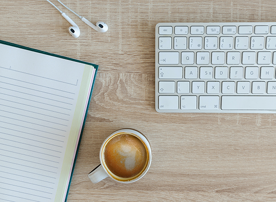 A notebook, coffee, headphones and keyboard sit on a desk