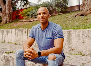 U of T Mississauga professor Akwasi Owusu-Bempah in a blue T-shirt and ripped jeans, sitting on a concrete boulder, behind which is a paved footpath with a short concrete wall and a grassy hill with trees on the other side