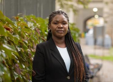 Ketty Anyeko stands outdoors in front of leafy bushes 